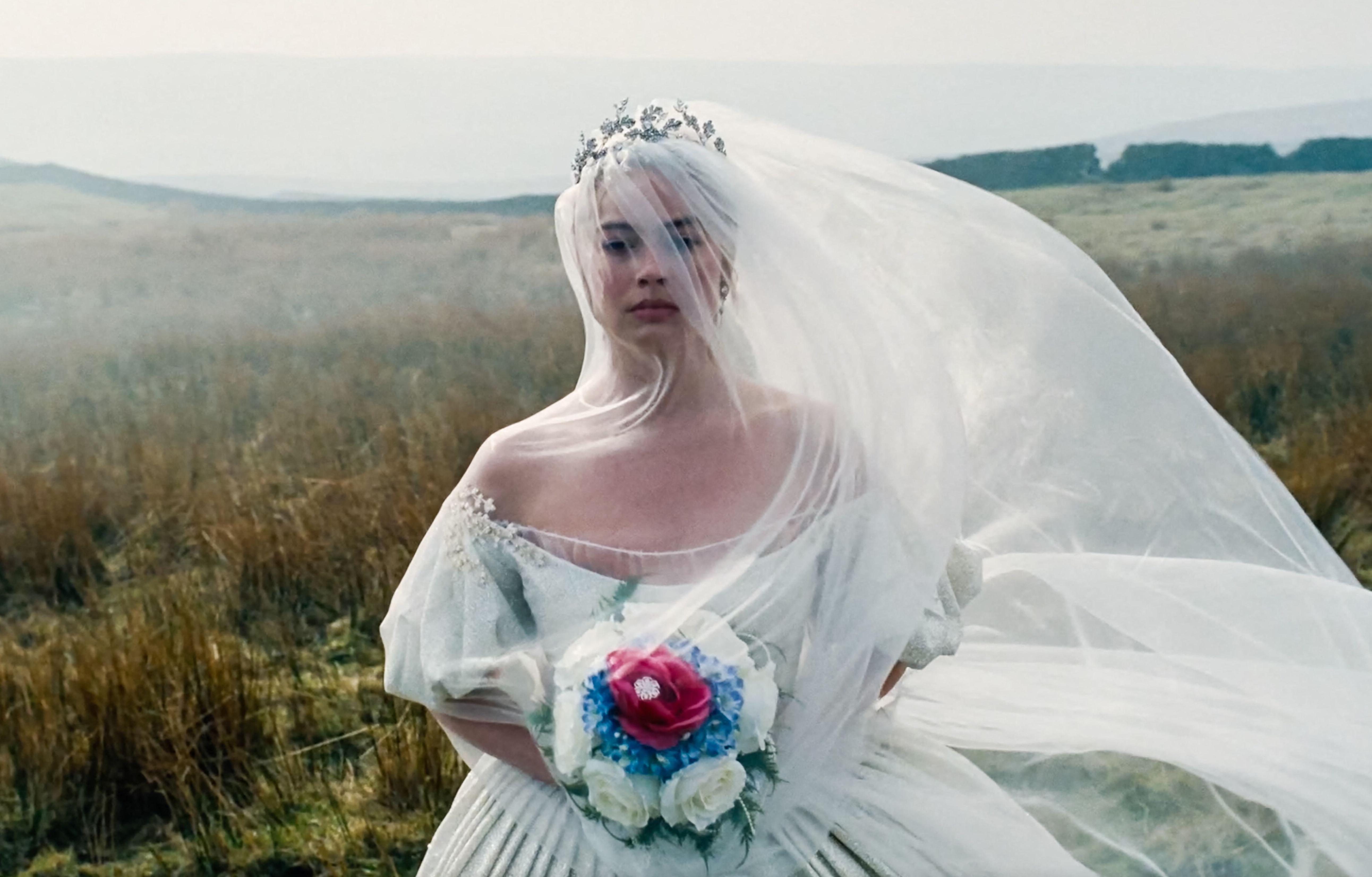 Margot Robbie as Catherine Earnshaw in a wedding dress and veil, holding a bouquet of flowers, standing in a field.