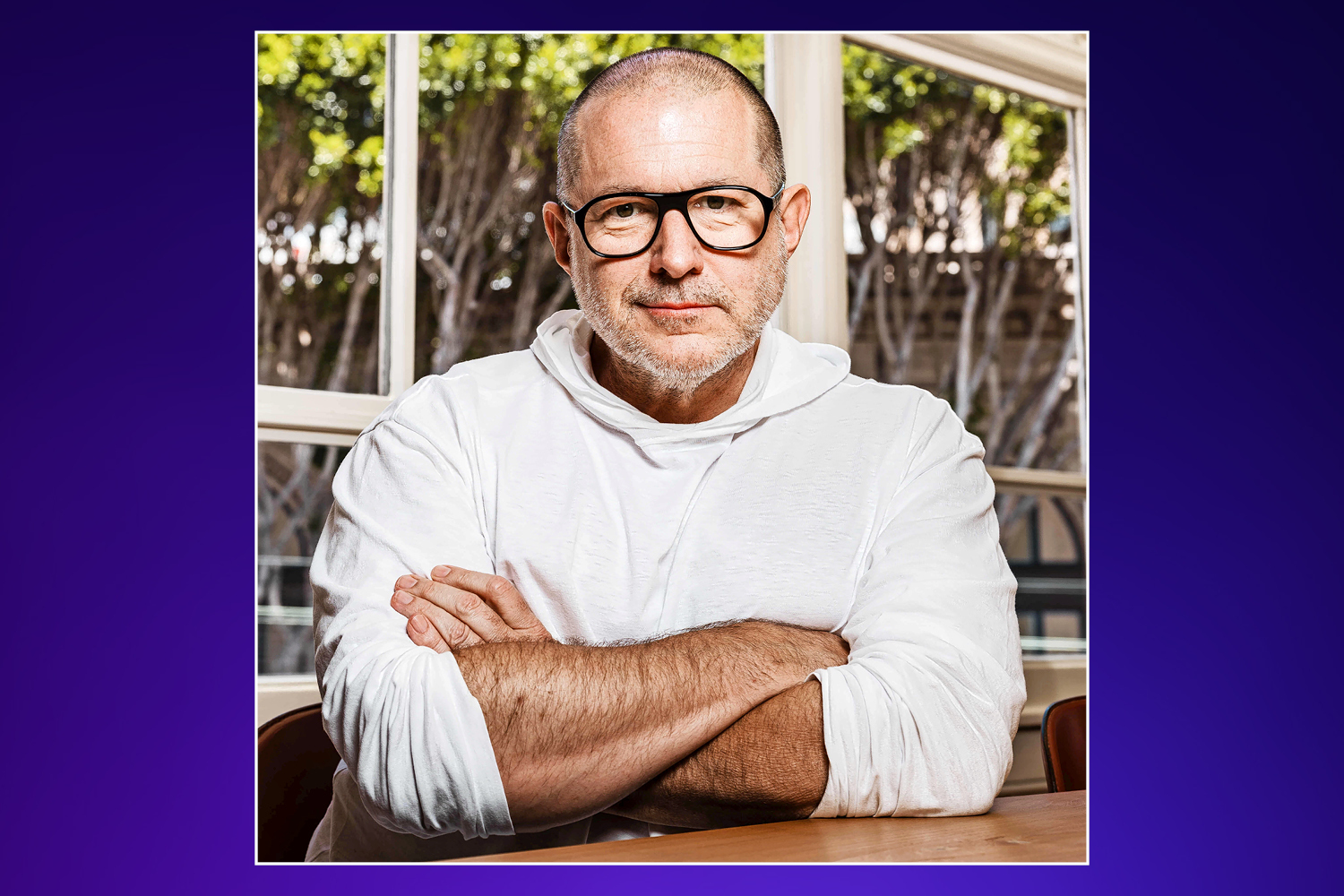 Man in white hooded shirt with arms crossed, wearing glasses, against a window with trees outside.