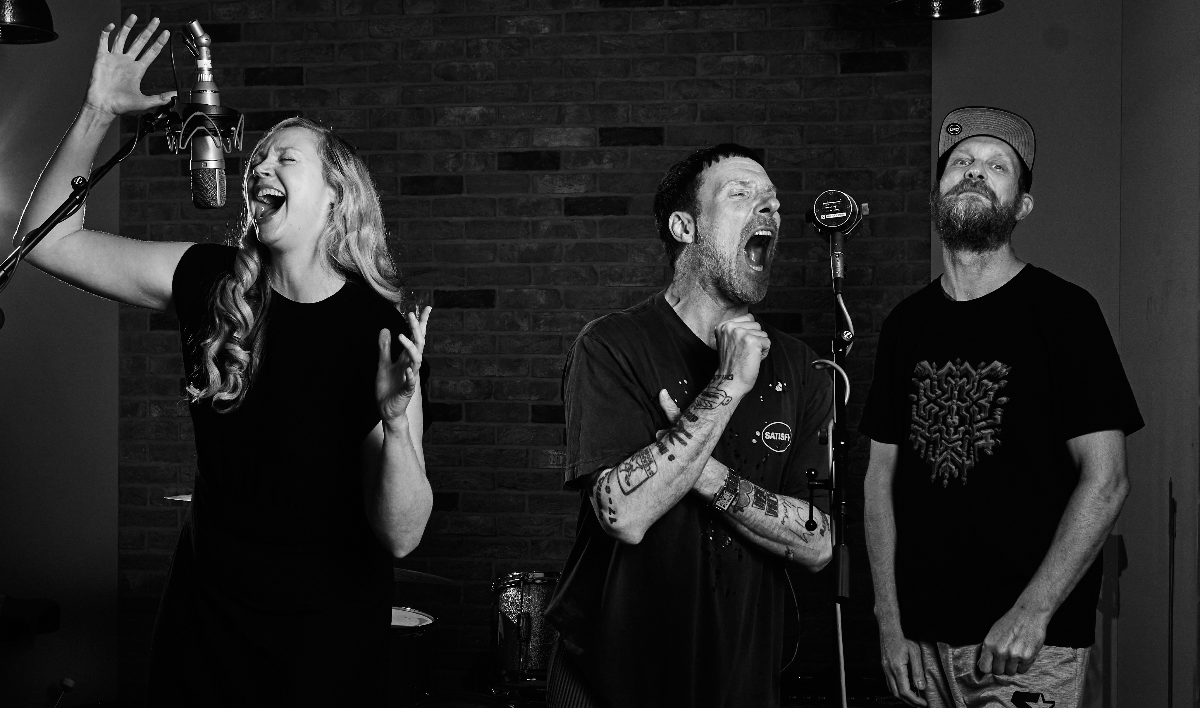 Gwendoline Christie singing into a microphone next to two members of Sleaford Mods in a recording studio.