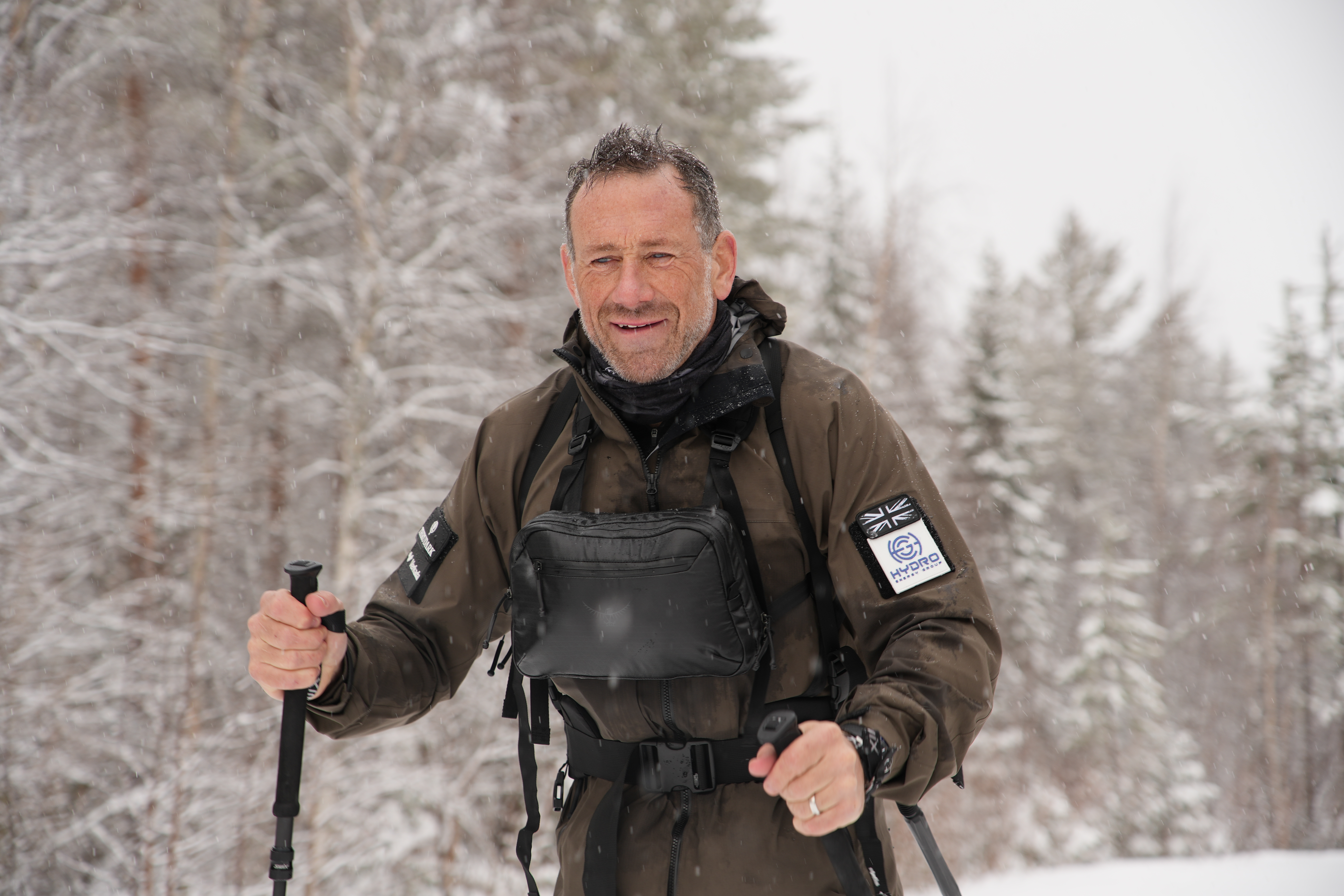 A man in a winter coat and hiking poles smiles at the camera, with a snowy forest in the background.