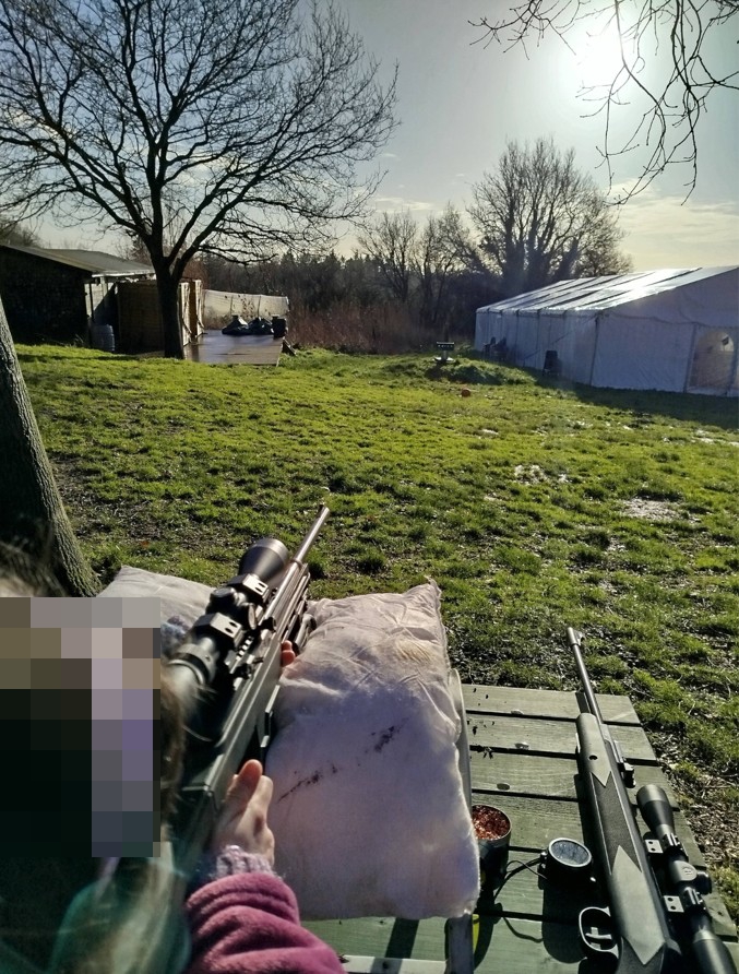 A child, with their face pixelated, aiming an air rifle from behind a wooden bench with a second rifle resting next to them.
