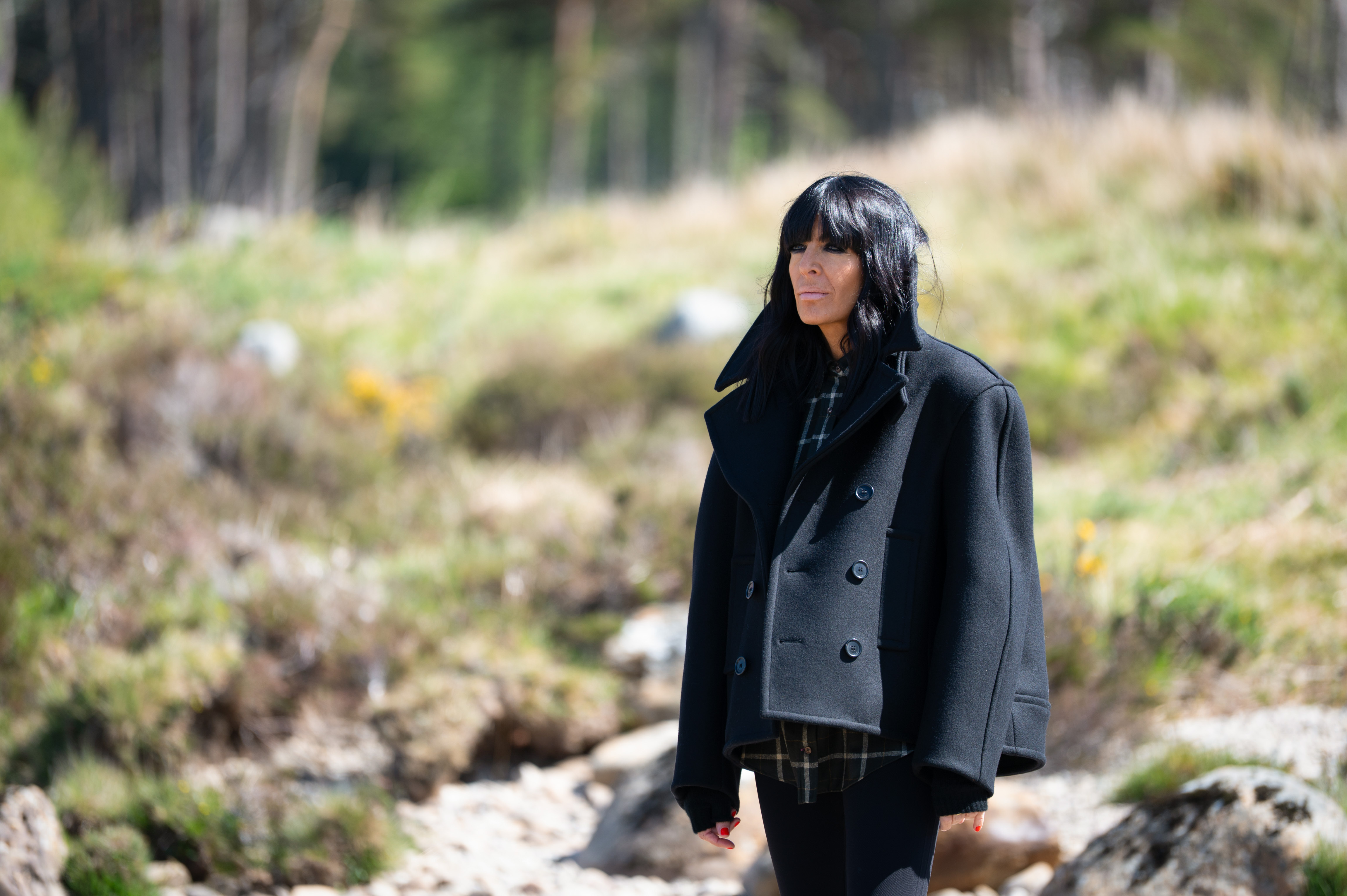 Claudia Winkleman, presenter of The Traitors, looking to the side outdoors, wearing a black coat over a plaid shirt.