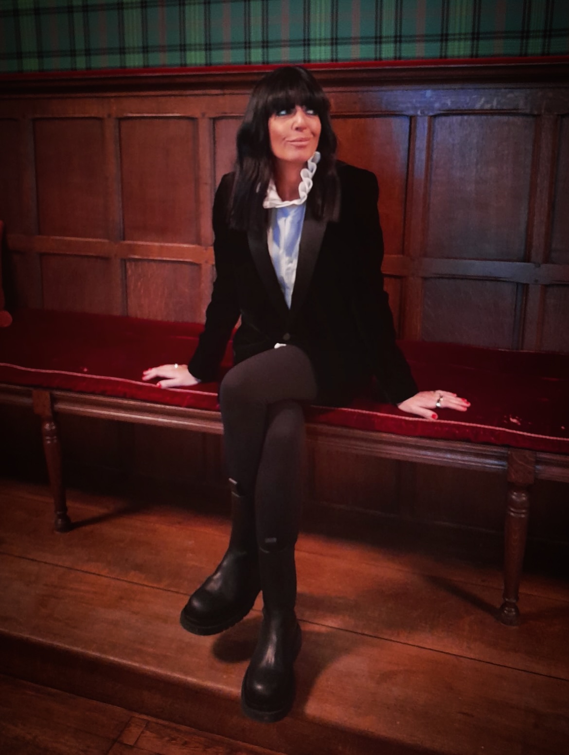 Claudia Winkleman on a red bench wearing a black velvet blazer and black boots.
