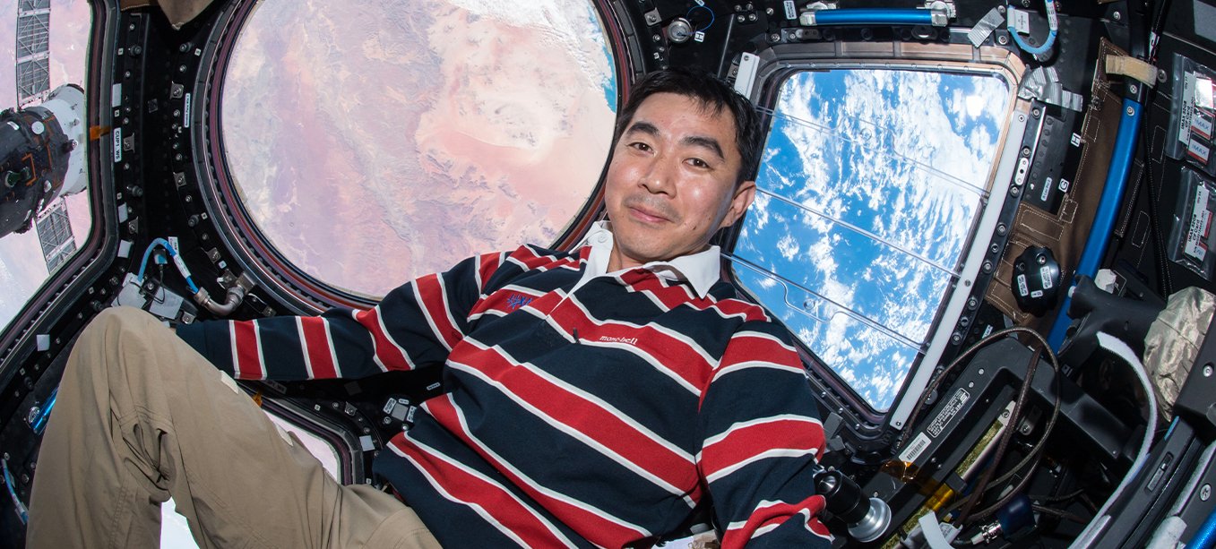 Astronaut Kimiya Yui aboard the International Space Station, smiling, with views of Earth from the cupola windows.
