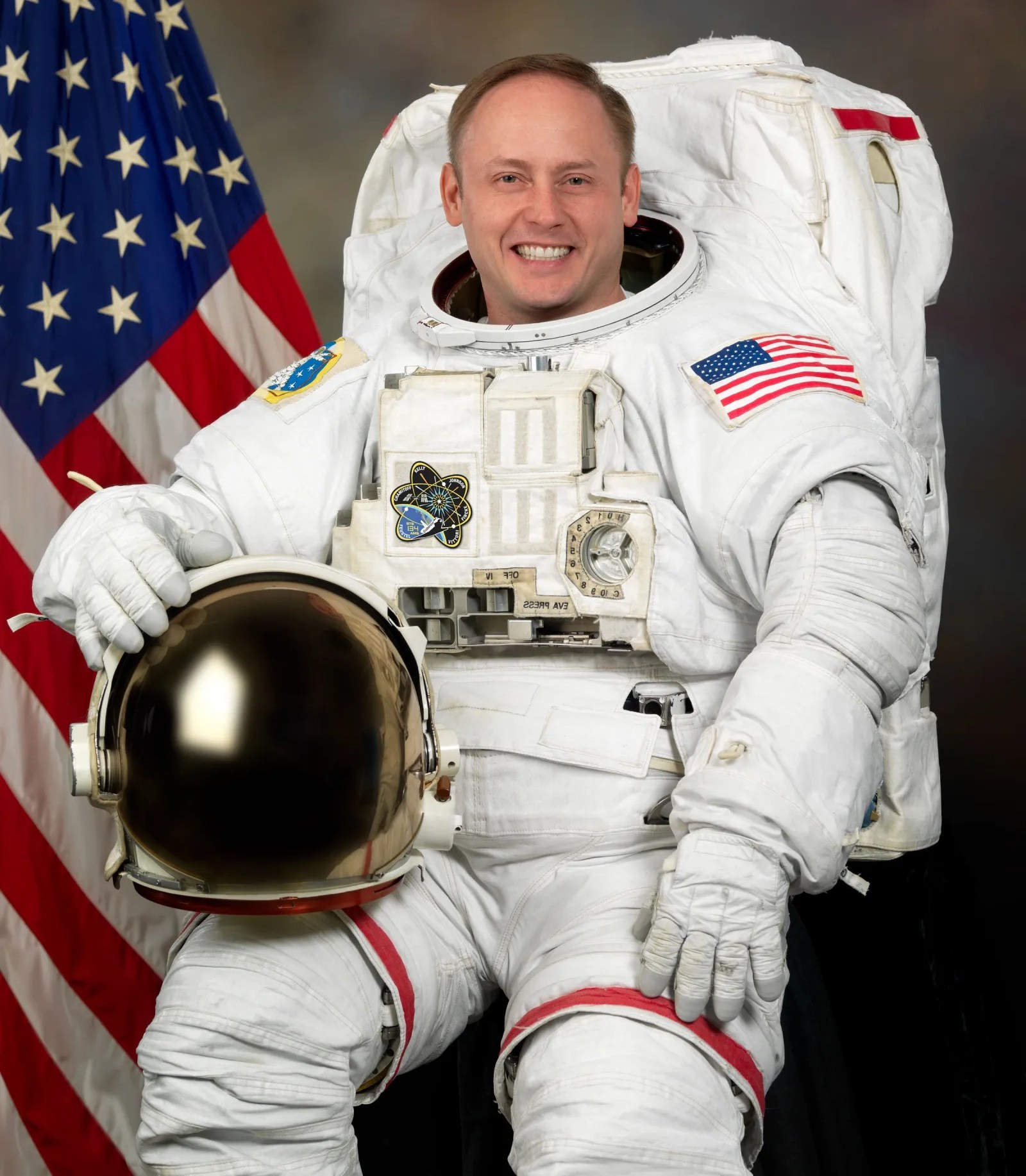 Astronaut Mike Fincke in a spacesuit holding a helmet, with an American flag in the background.