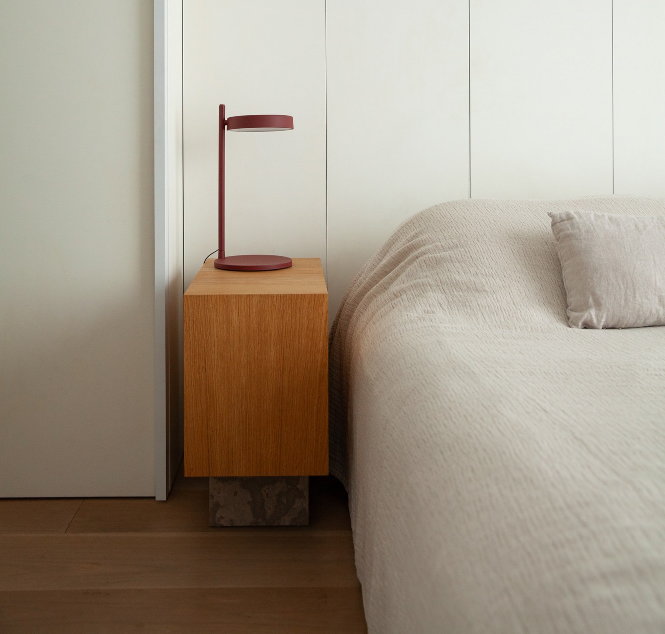 Bedroom interior with a red lamp on a wooden nightstand next to a bed with a light-colored duvet.