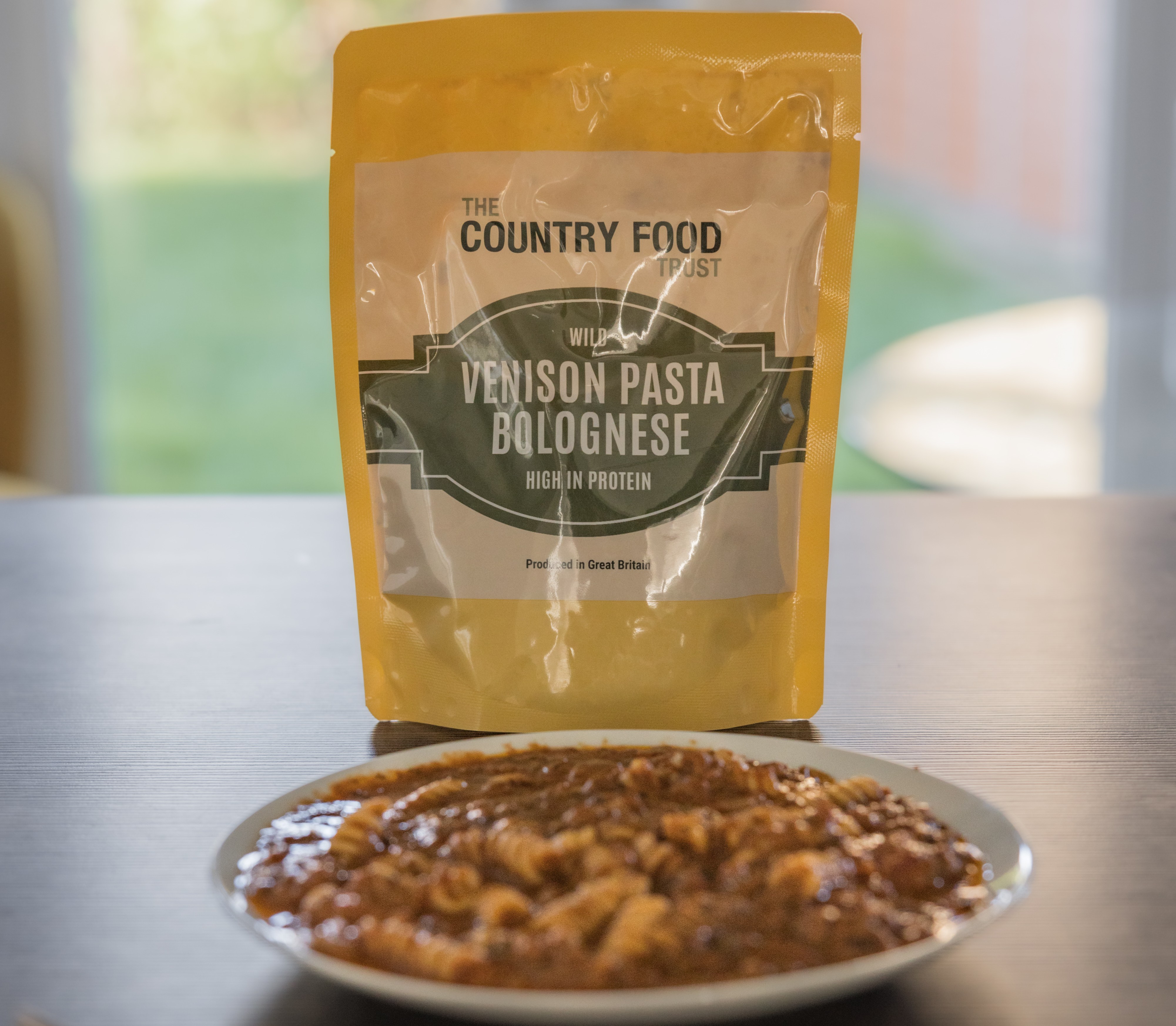 A package of "The Country Food Trust Wild Venison Pasta Bolognese, High in Protein" next to a plate of cooked venison pasta Bolognese and a fork.