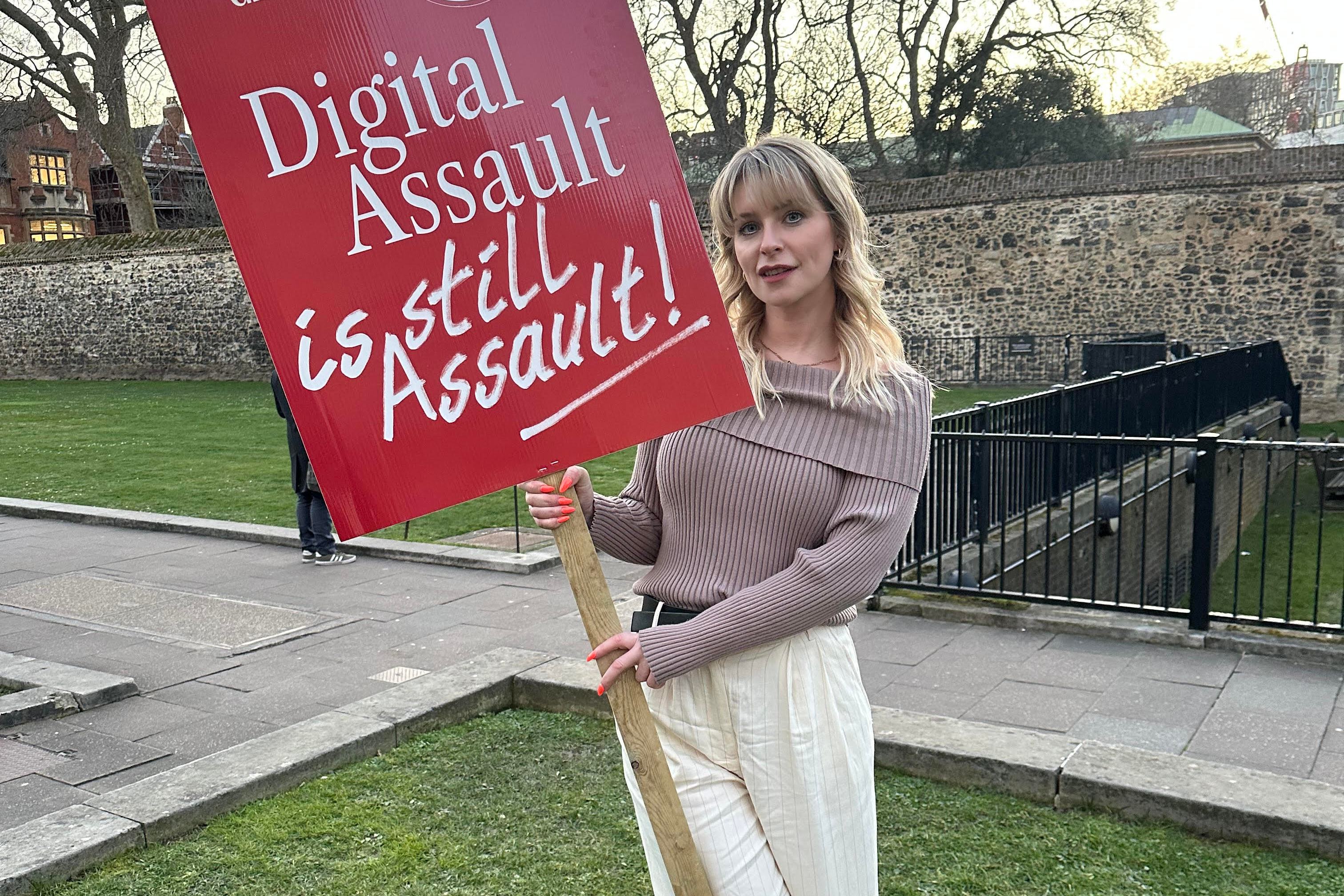 Jess Davies, an author and campaigner, holds a red protest sign that reads "Digital Assault is Still Assault!" outside.