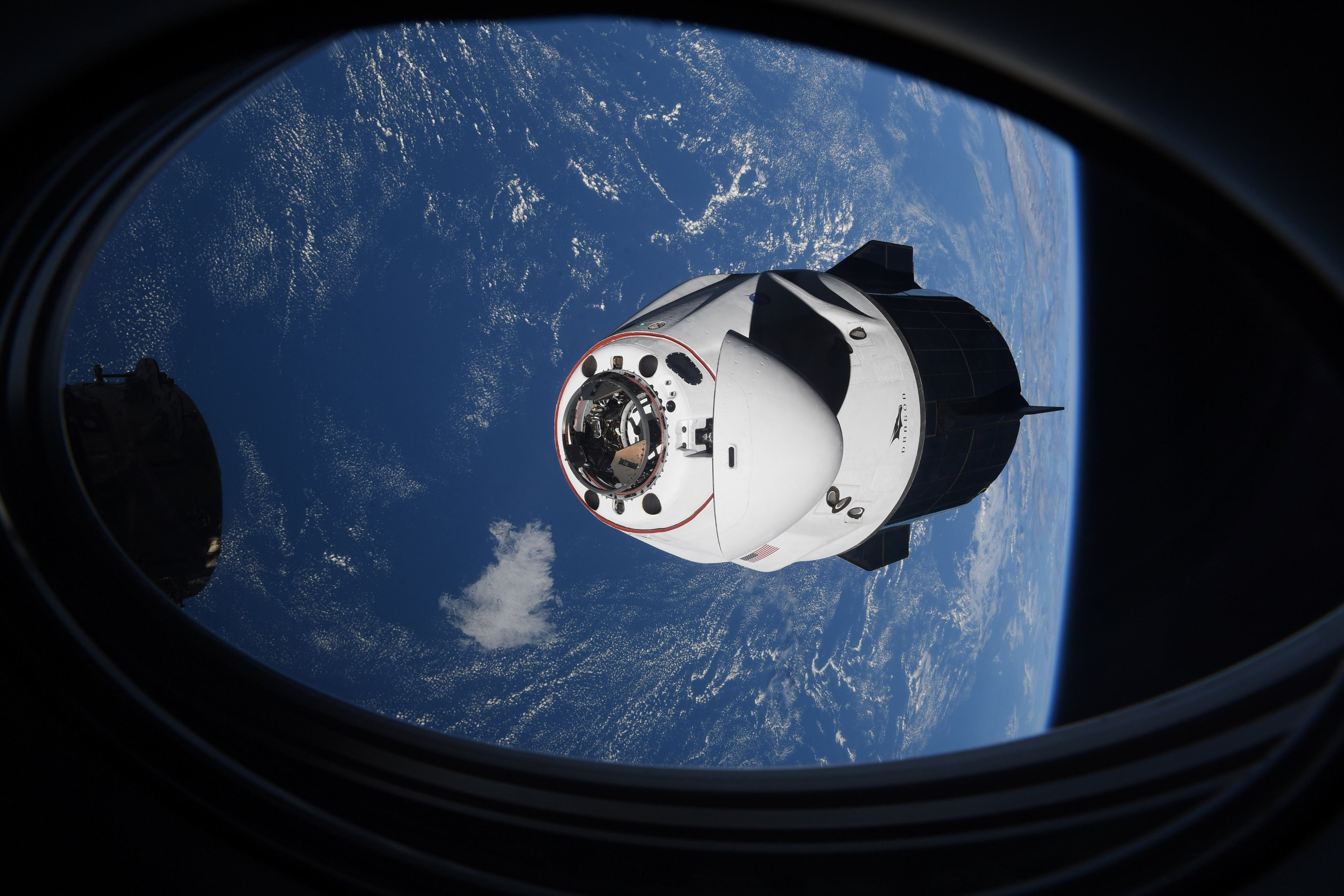 A SpaceX Crew Dragon capsule approaches the International Space Station against the blue Earth below, as seen through a spacecraft window.