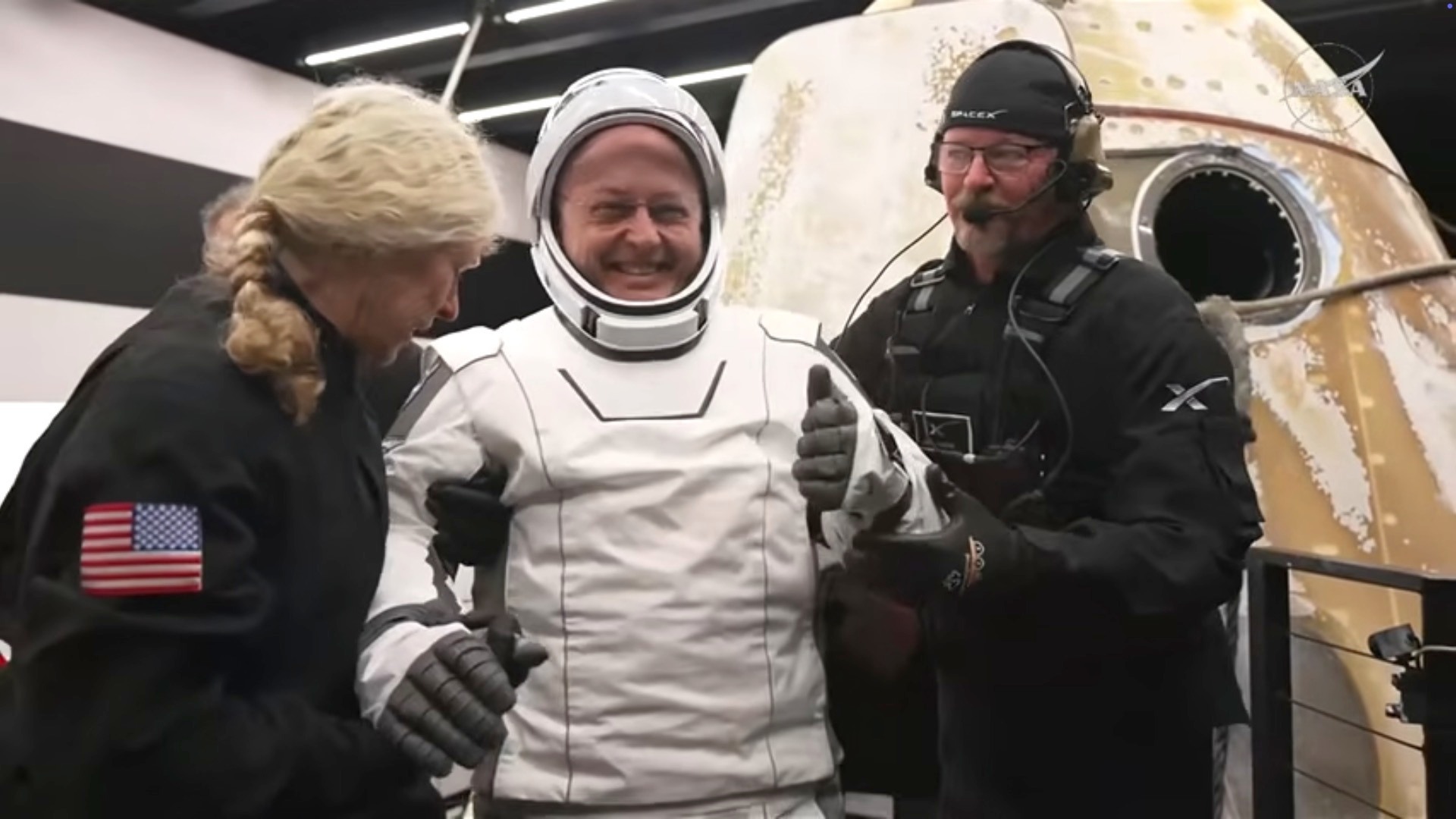 Astronaut Mike Fincke in a white space suit smiles and gives a thumbs-up as he is helped out of the SpaceX Crew-11 capsule by two people.
