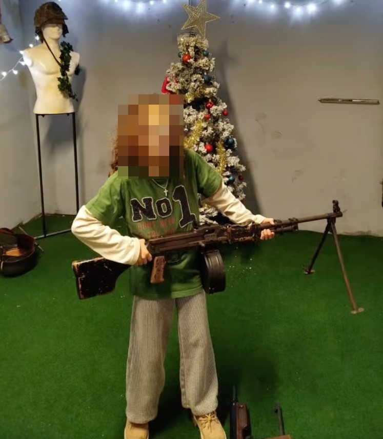 A child holding a large machine gun in front of a decorated Christmas tree.