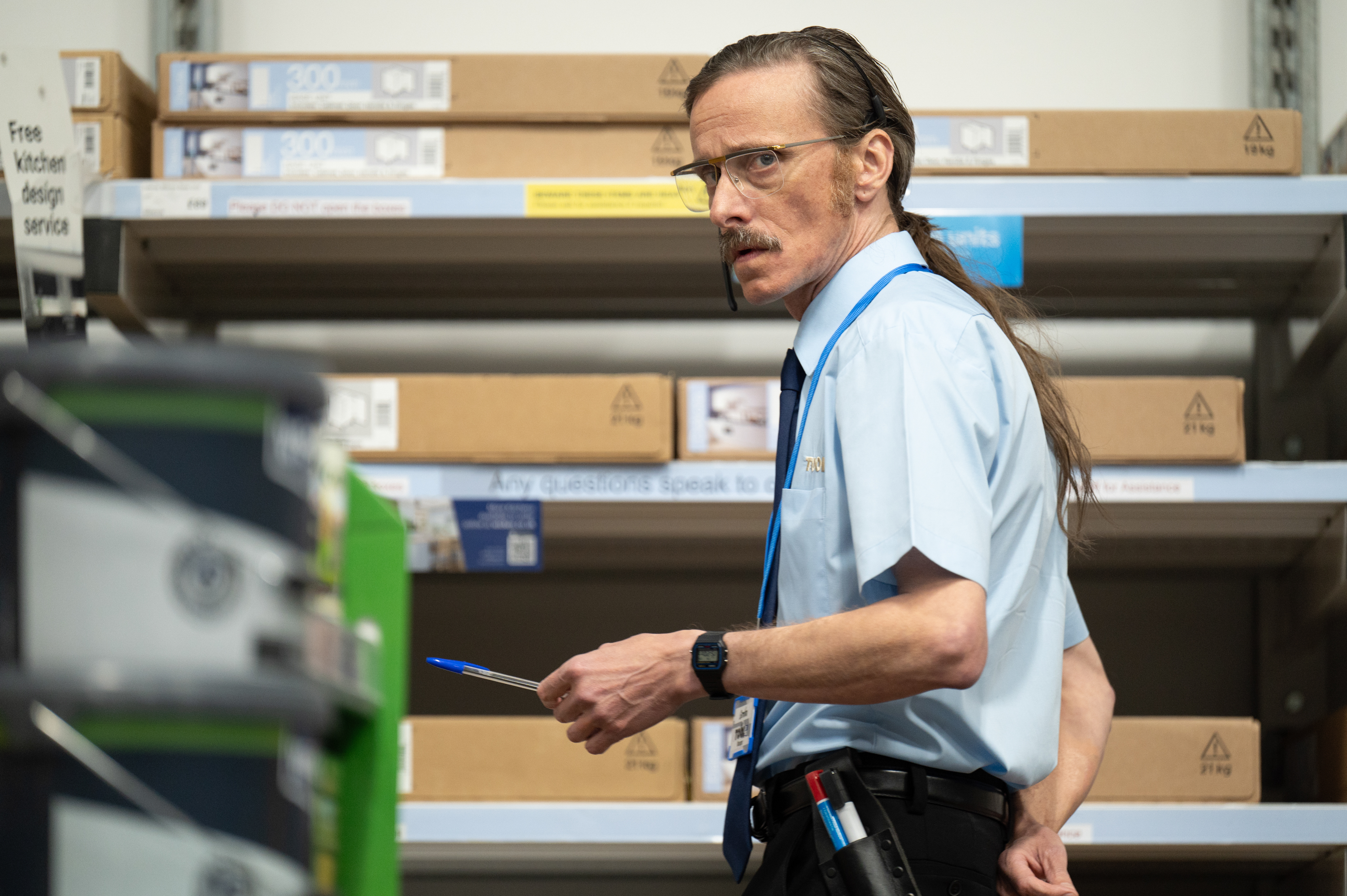 Mackenzie Crook as Gordon, a man wearing glasses, mustache, and a ponytail, holding a pen in a store aisle.
