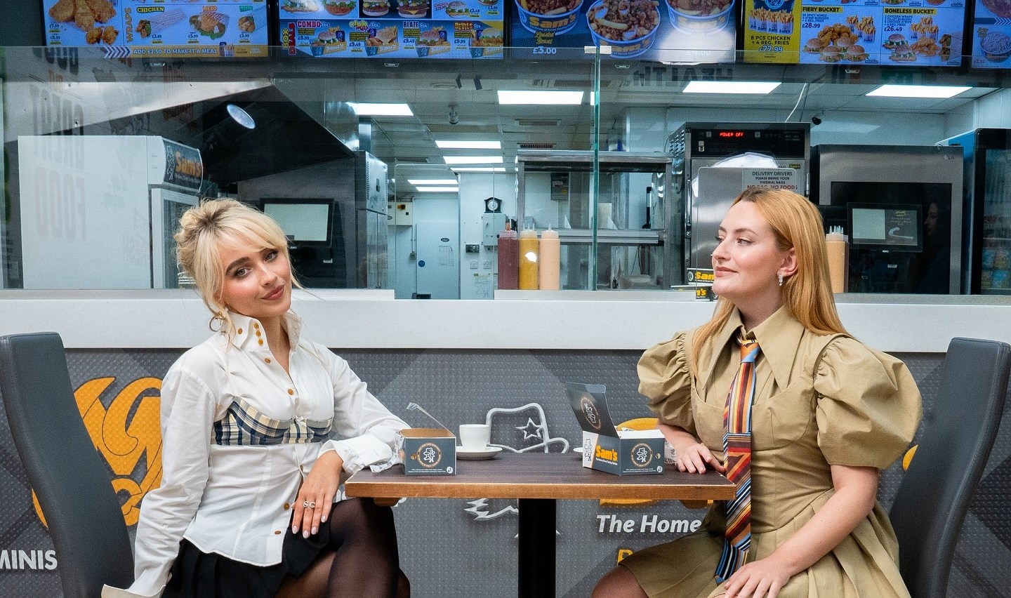 Two women sit at a table in a chicken shop, one wearing a white shirt and black skirt, the other a tan dress and striped tie.