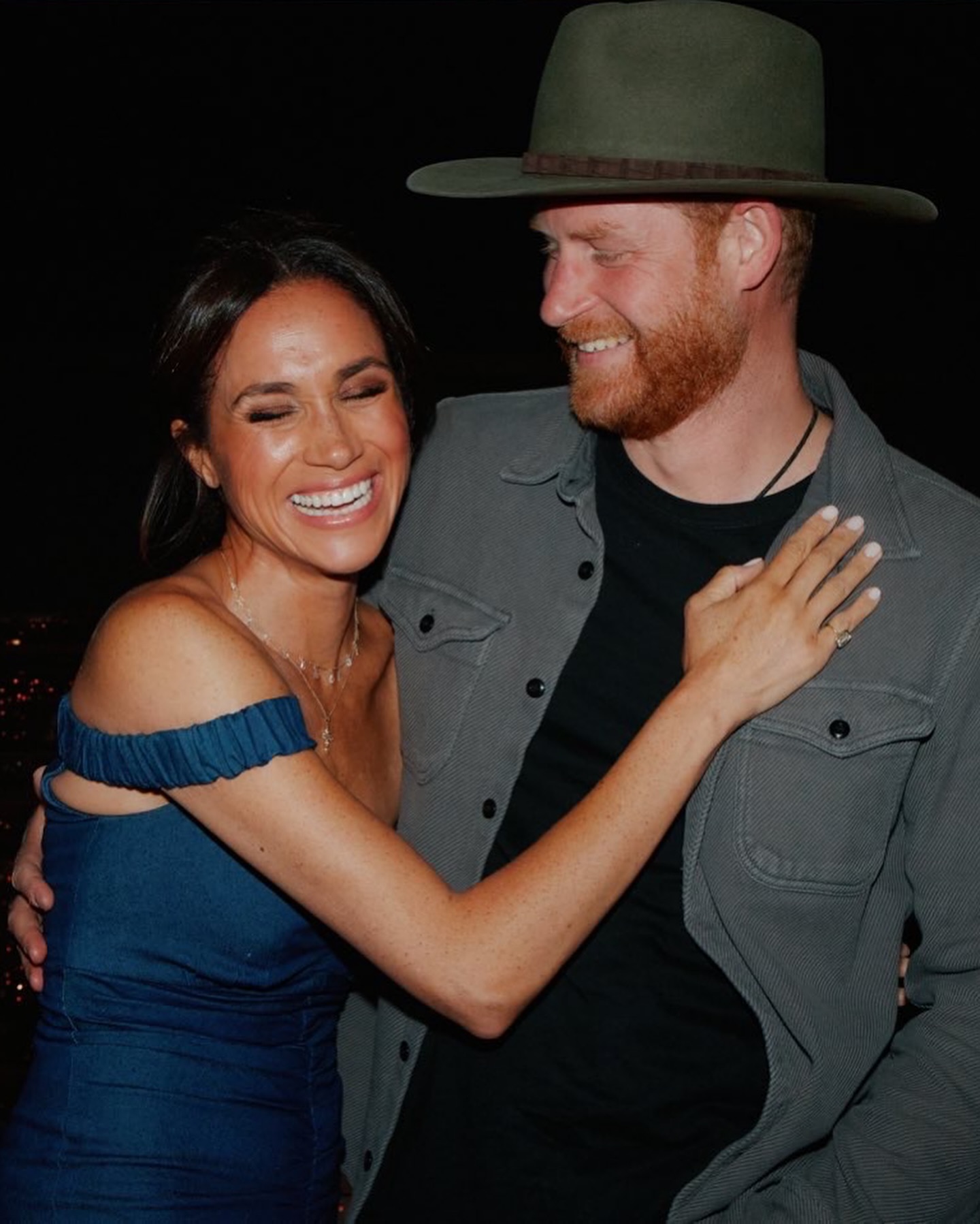 Meghan Markle smiling while embracing Prince Harry, who wears a hat.