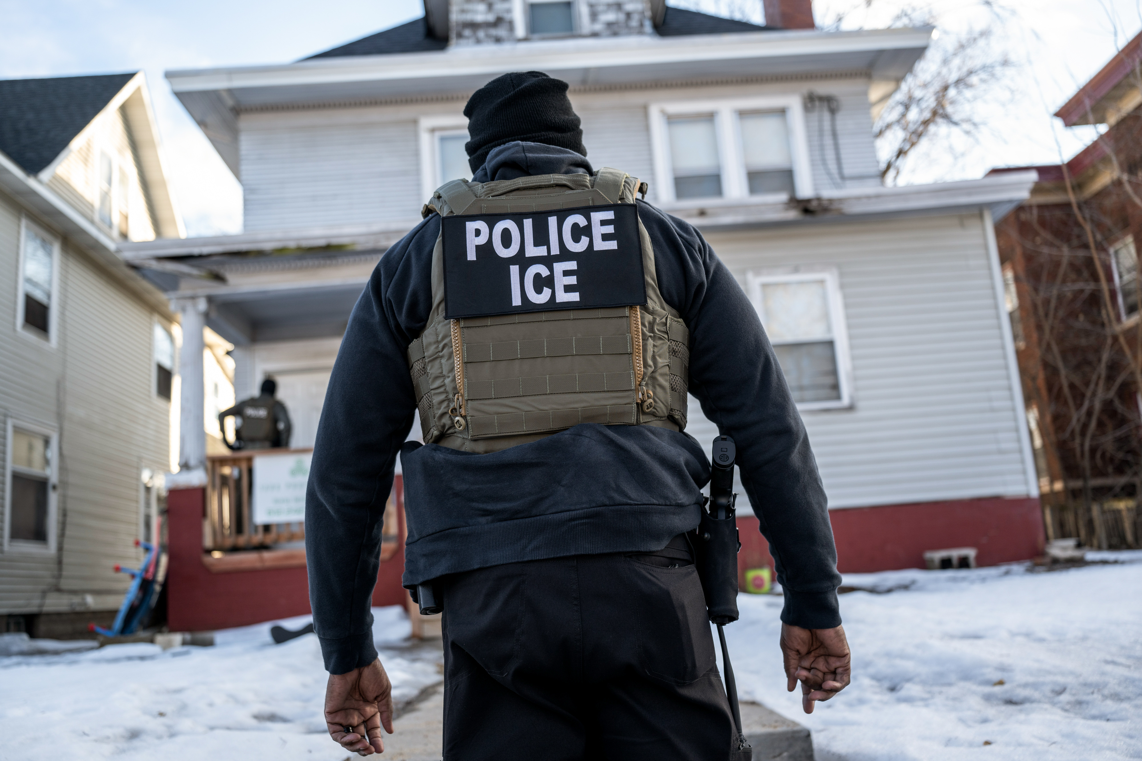 A federal ICE agent in a tactical vest with "POLICE ICE" on the back stands outside a house in Minneapolis.