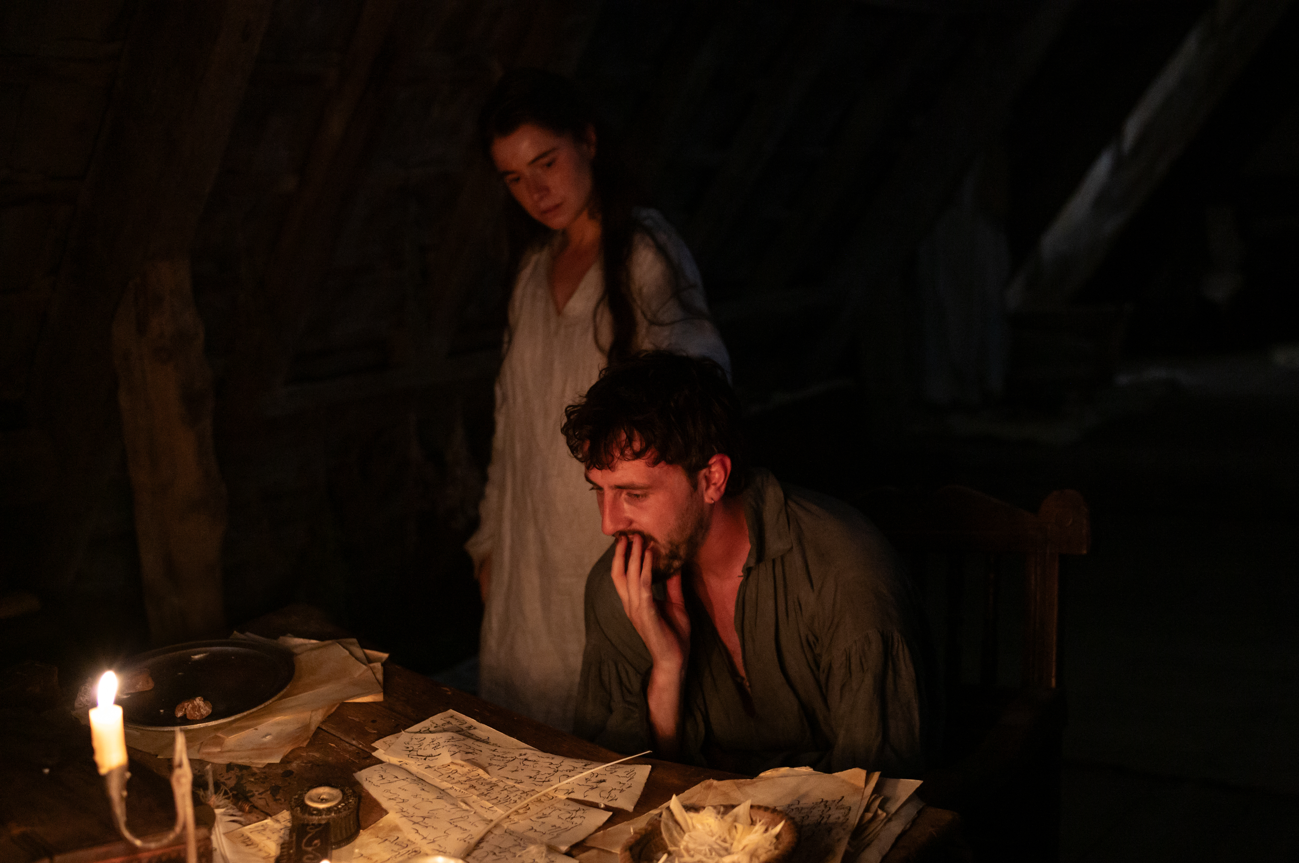 Jessie Buckley as Agnes standing behind Paul Mescal as William Shakespeare, who is seated at a table with papers and a quill, lit by candlelight.