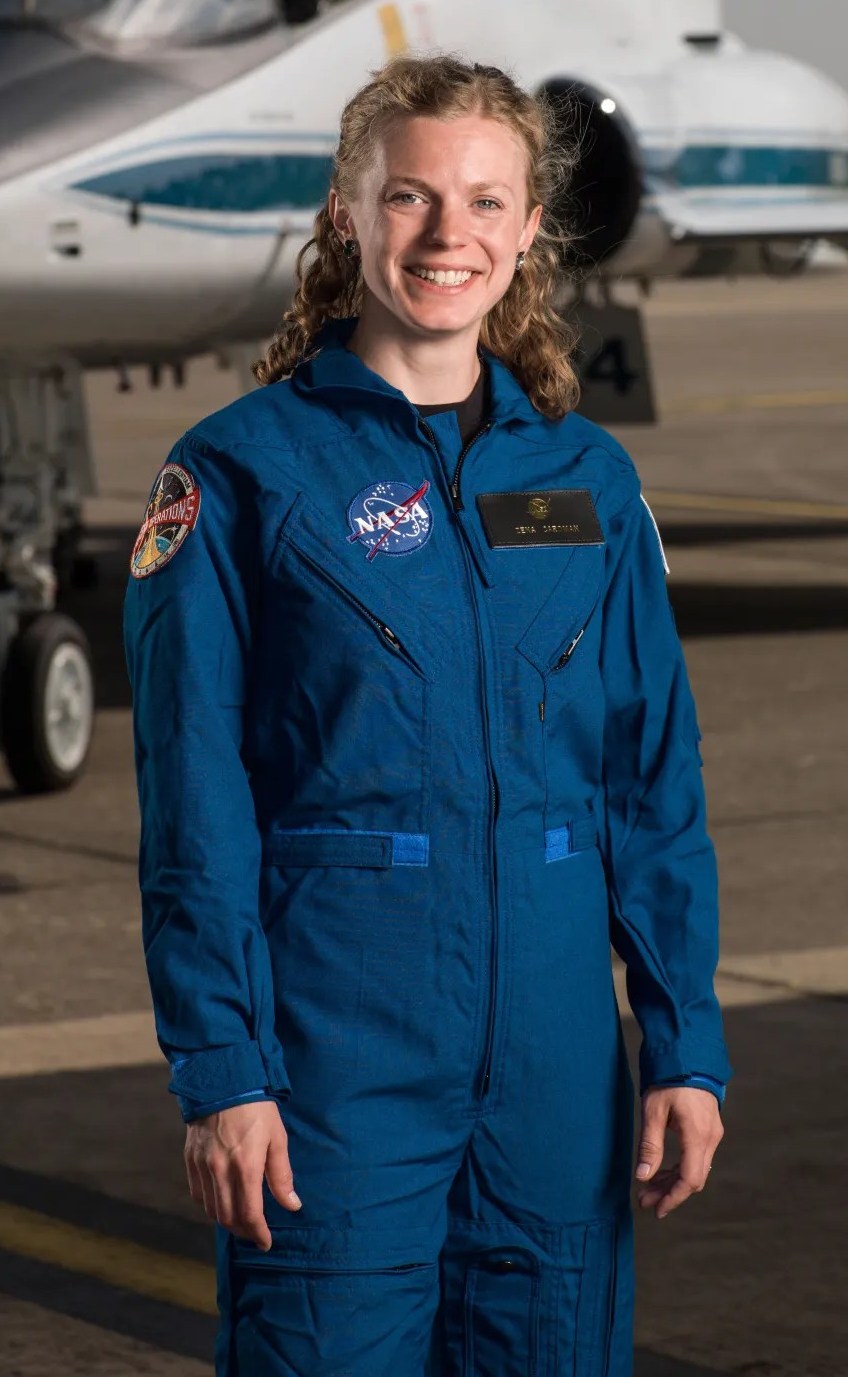 Astronaut Zena Cardman in a blue flight suit smiling in front of a white jet with its canopy open.