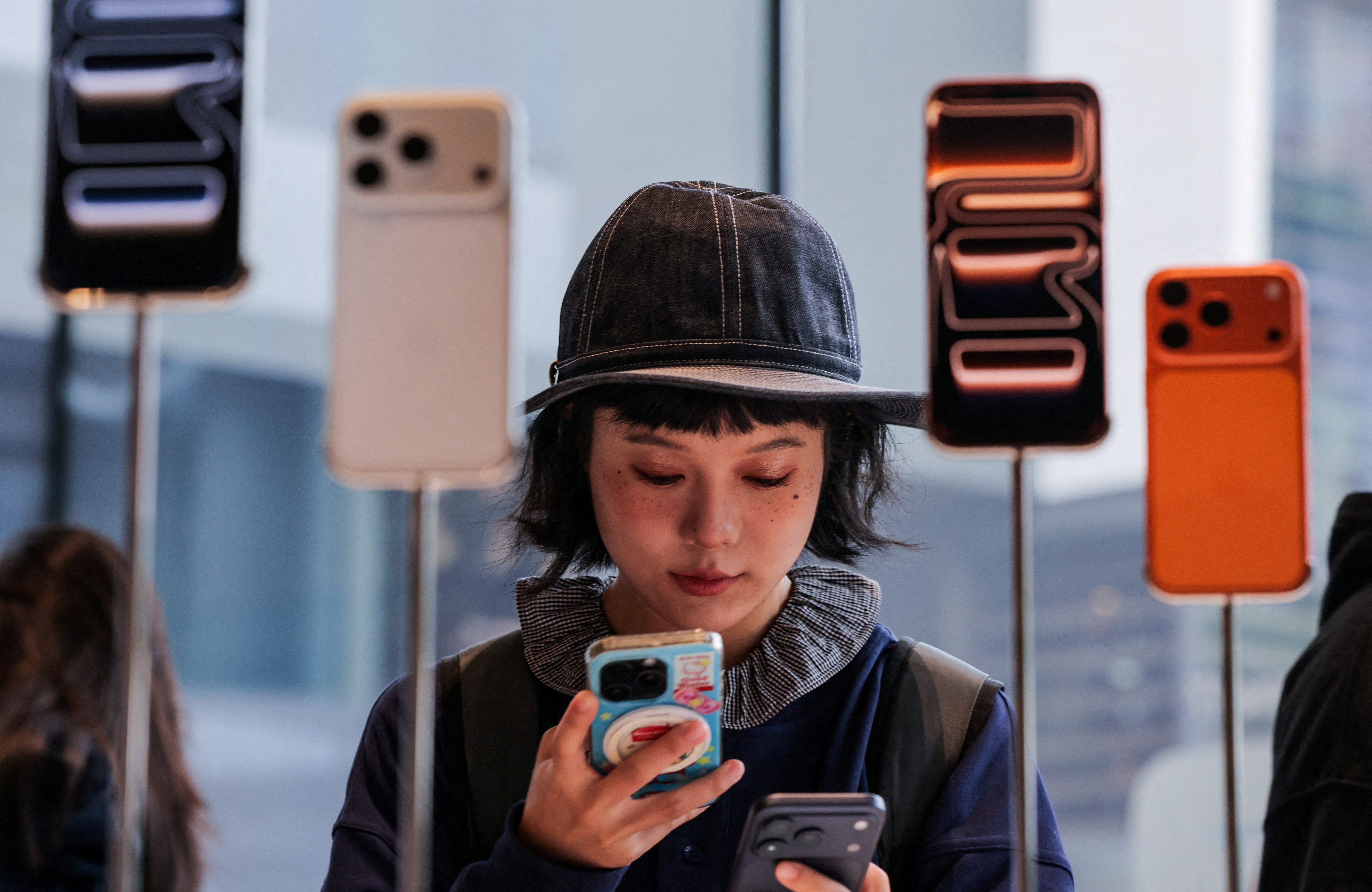 A woman in a hat uses her smartphone inside an Apple store with iPhone 17 series smartphones on display.