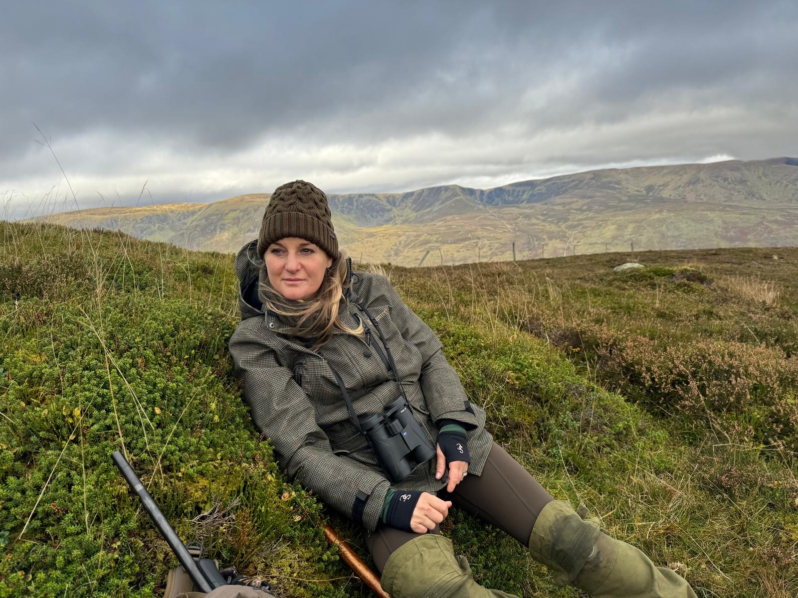 Deer stalker Jodie Simpson lying on a grassy hill with a rifle and binoculars, with mountains in the background.