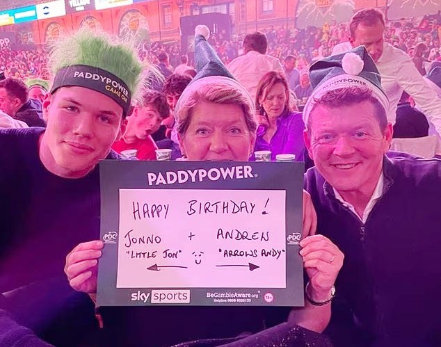 Jonno (left), his father Andrew (right), and BBC presenter Clare (center) at a darts event, holding a sign that reads "Happy Birthday! Jonno + Andrew".