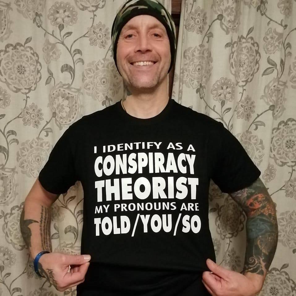 A man wearing a black t-shirt that reads "I IDENTIFY AS A CONSPIRACY THEORIST MY PRONOUNS ARE TOLD/YOU/SO".