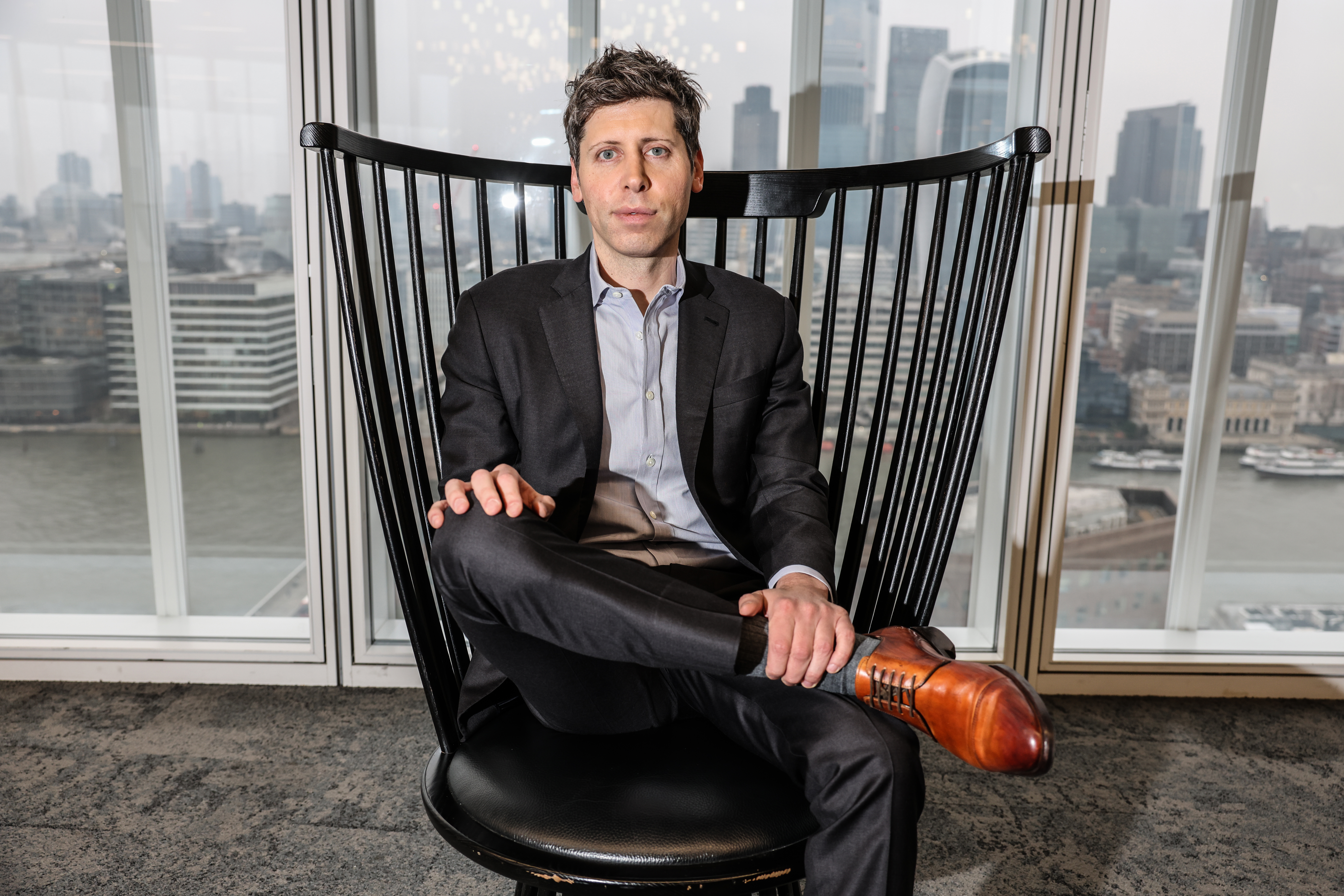 Sam Altman, founder of Open AI, sits in a chair with his legs crossed in front of a window overlooking a cityscape.