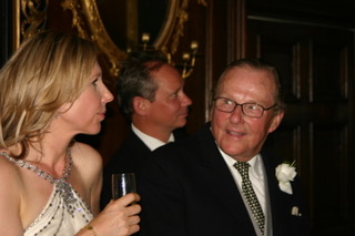 A woman in a beaded dress holding a champagne flute smiles while looking at an older man in a suit and tie.