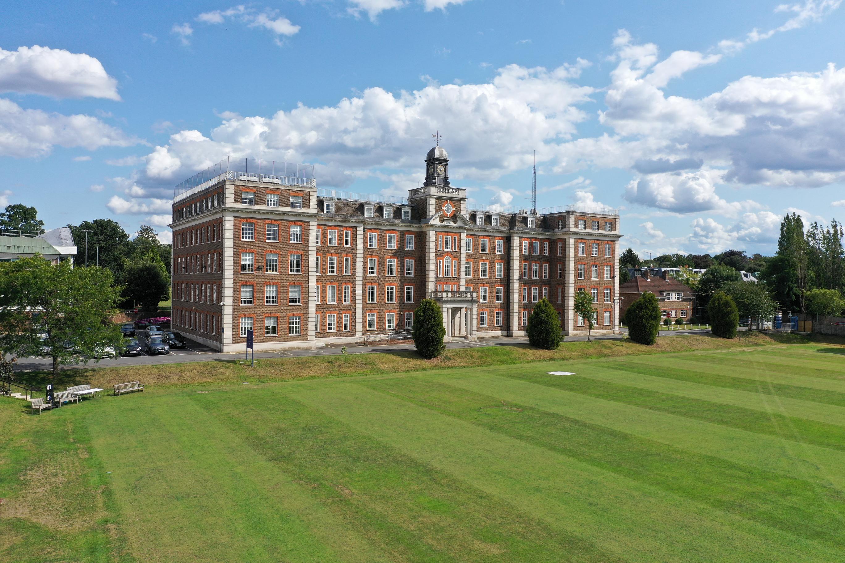 An aerial view of the Bank of England's Roehampton sports ground and former records office, a large brick building with a clock tower next to a manicured lawn.
