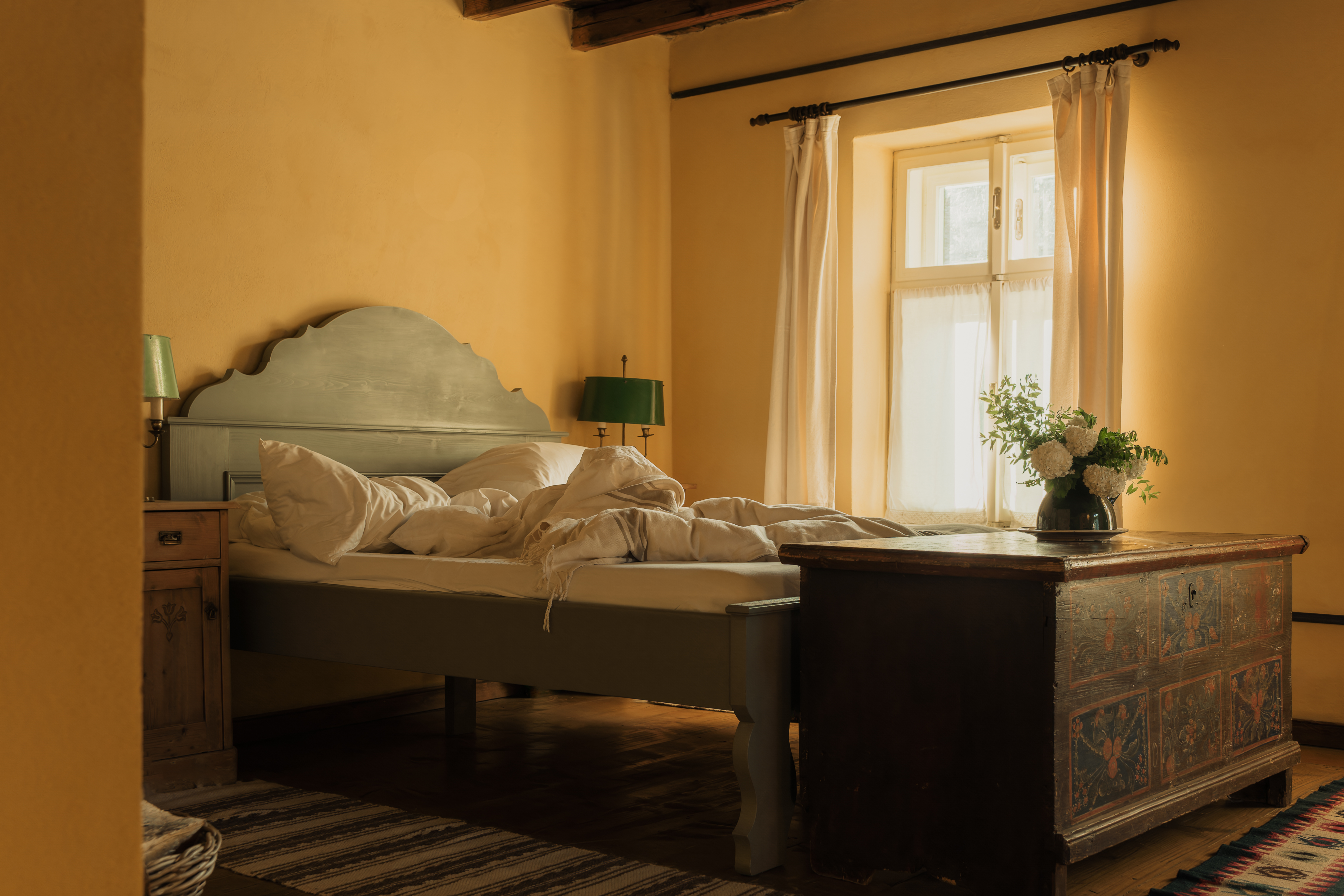 A bedroom with a bed with white bedding, a window with white curtains, a green lamp, a wooden bedside table, and a decorative wooden trunk with white flowers.