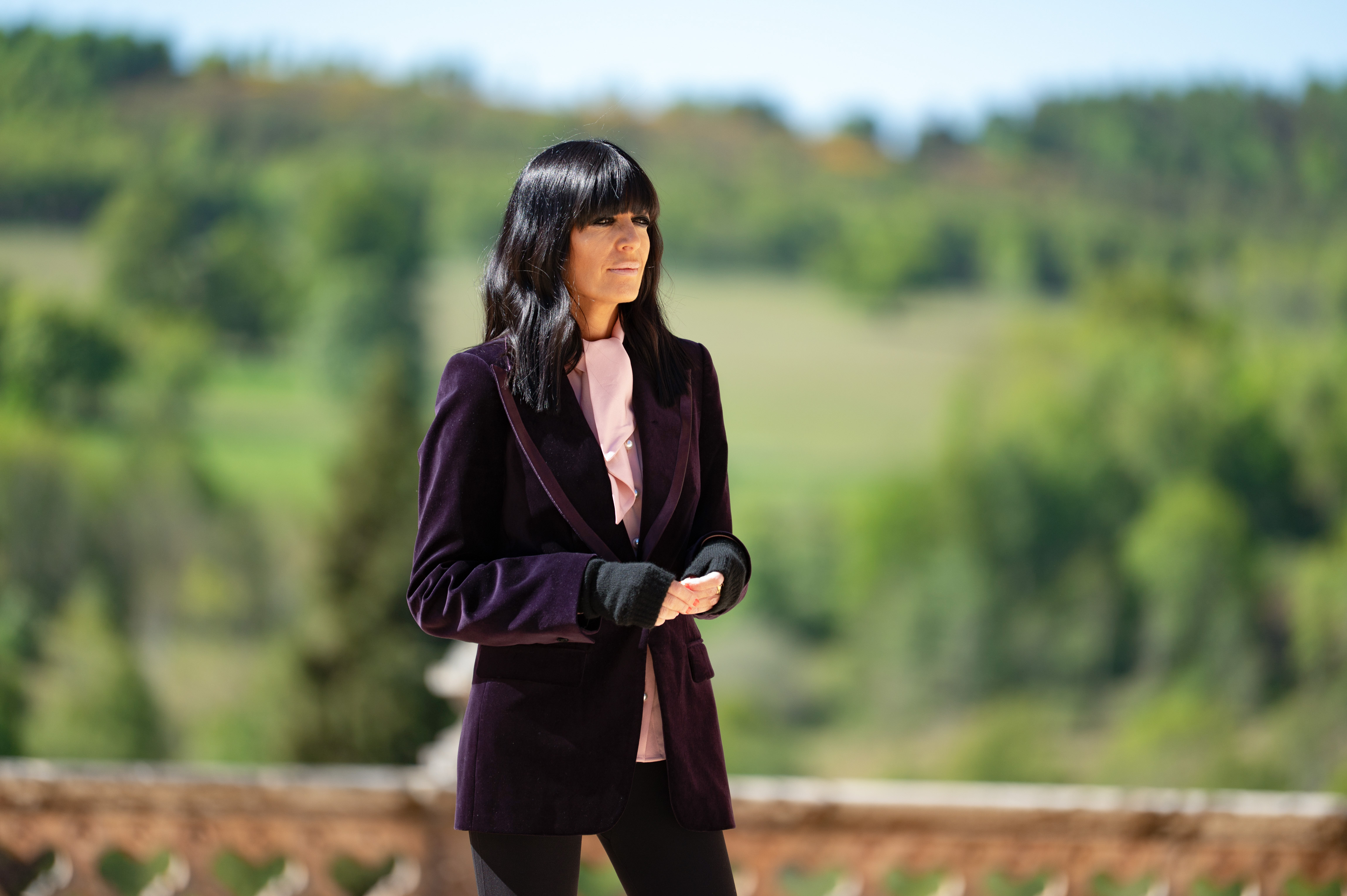 Claudia Winkleman, wearing a purple velvet jacket and pink shirt, stands outdoors.