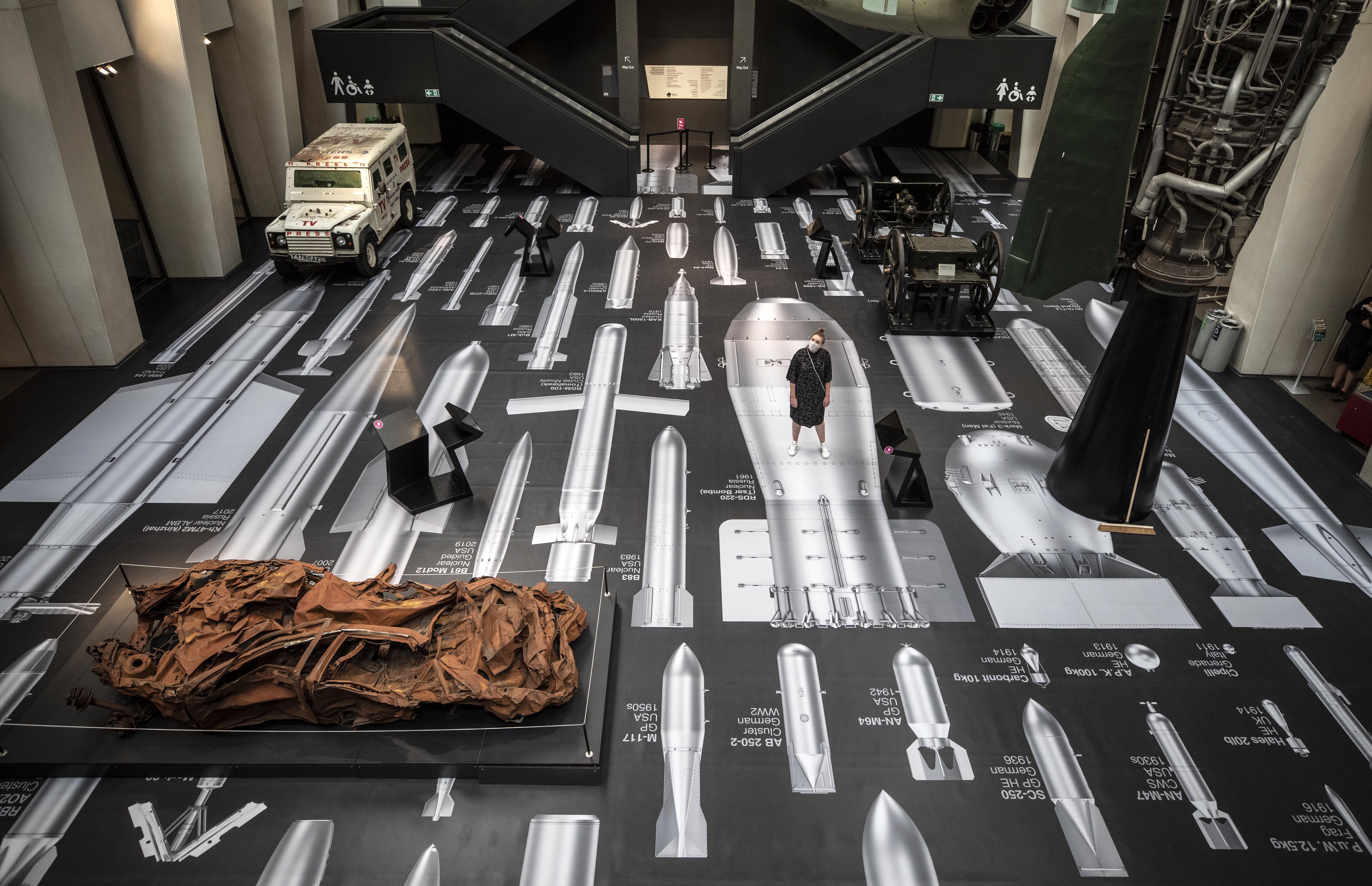 Ai Weiwei's "History of Bombs" installation at the Imperial War Museum, featuring bomb illustrations on the floor and walls, and a crushed vehicle.