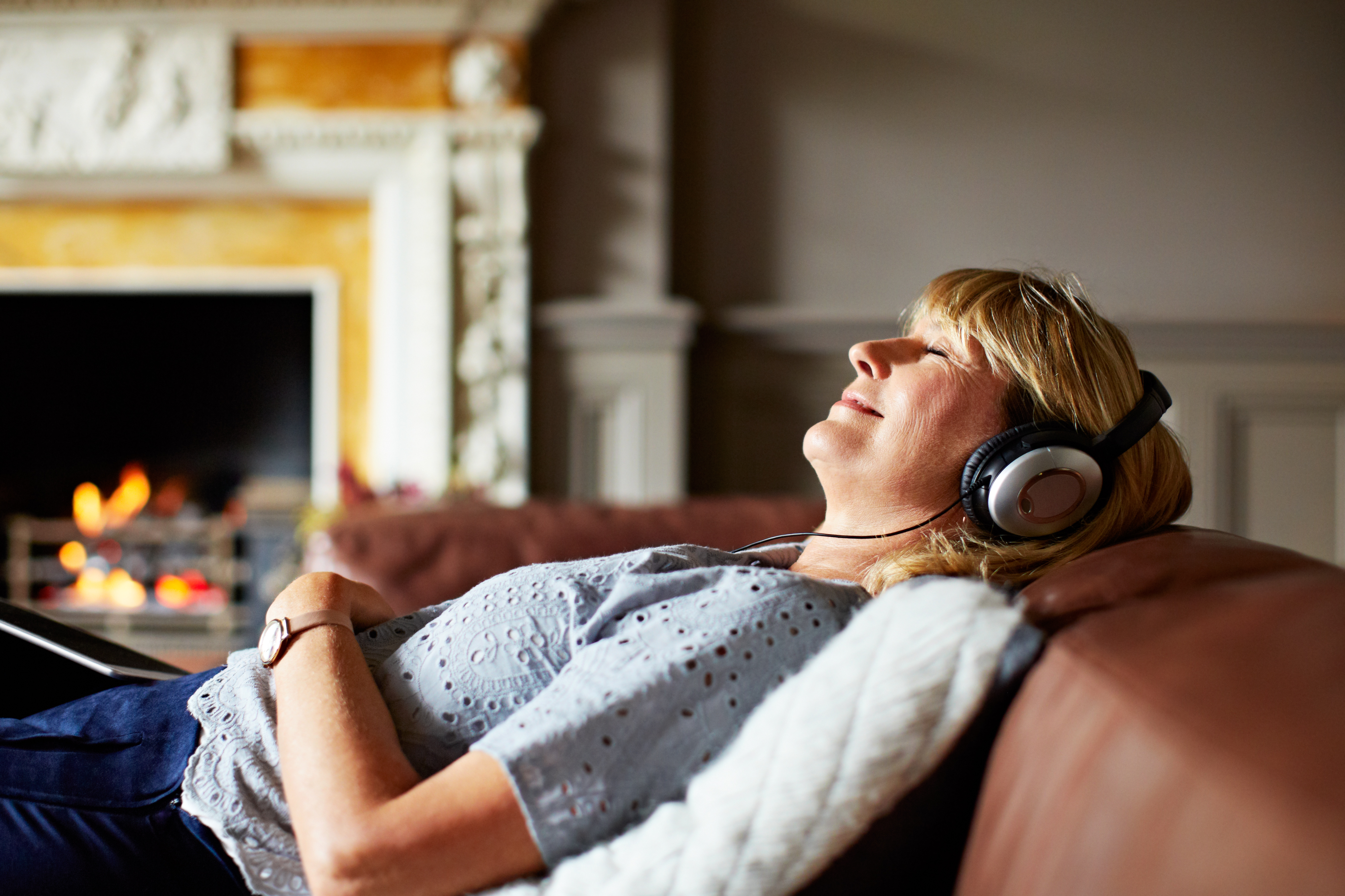 A mature woman with blonde hair, wearing headphones, lying on a couch with her eyes closed, listening to music with a fireplace in the background.