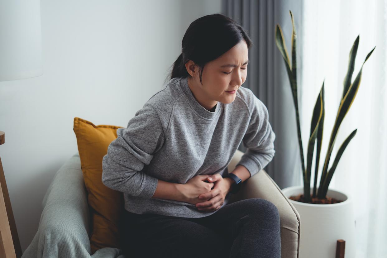 Asian woman feeling stomachache and touching her belly sitting on armchair in living room at home.