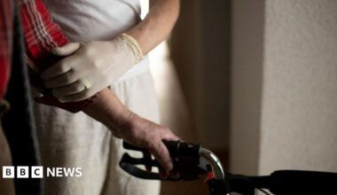 The arm of an elderly person holding onto to a walking aid. His arm is being supported by a person wearing a latex glove.