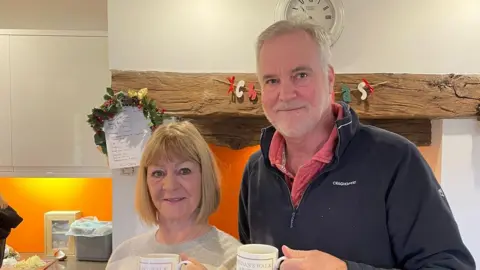 Brendan Wilson A cosy kitchen scene, Celia and Brendan are standing side by side, holding matching white mugs with printed text on them. They are positioned in front of a large wooden beam and an inglenook‑style fireplace adorned with a small festive wreath and decorative letters spelling “Christmas.” Behind them are clean white cabinets and a lit orange wall. The atmosphere is warm and homely.