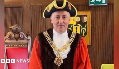 A false image of Ashley Bowkett the mayor of Gloucester in his mayoral robes ceremonial chain and traditional hat. The fake image depicts him standing in the council chamber with a large old wooden traditional chair behind him with a red leather back and a wooden panelled wall.