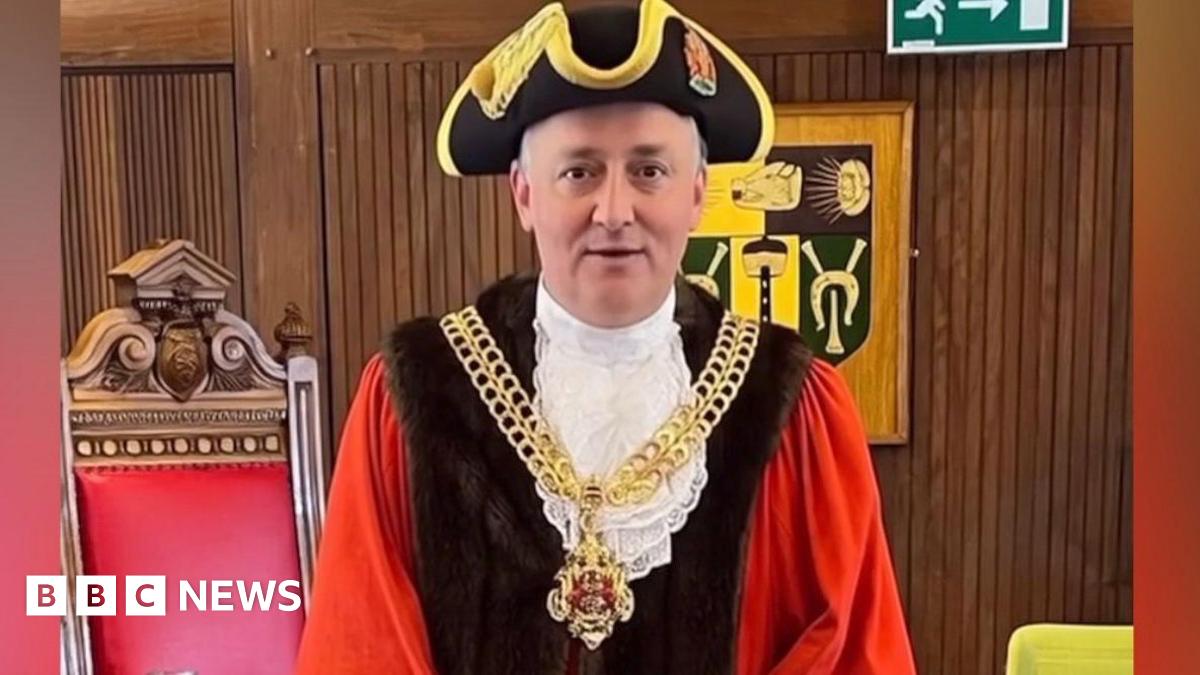 A false image of Ashley Bowkett the mayor of Gloucester in his mayoral robes ceremonial chain and traditional hat. The fake image depicts him standing in the council chamber with a large old wooden traditional chair behind him with a red leather back and a wooden panelled wall.