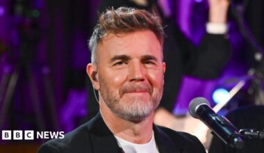Headshot of Barlow in front of a mic. He wears an in-ear monitor. You can see the top of his white t-shirt and black blazer.