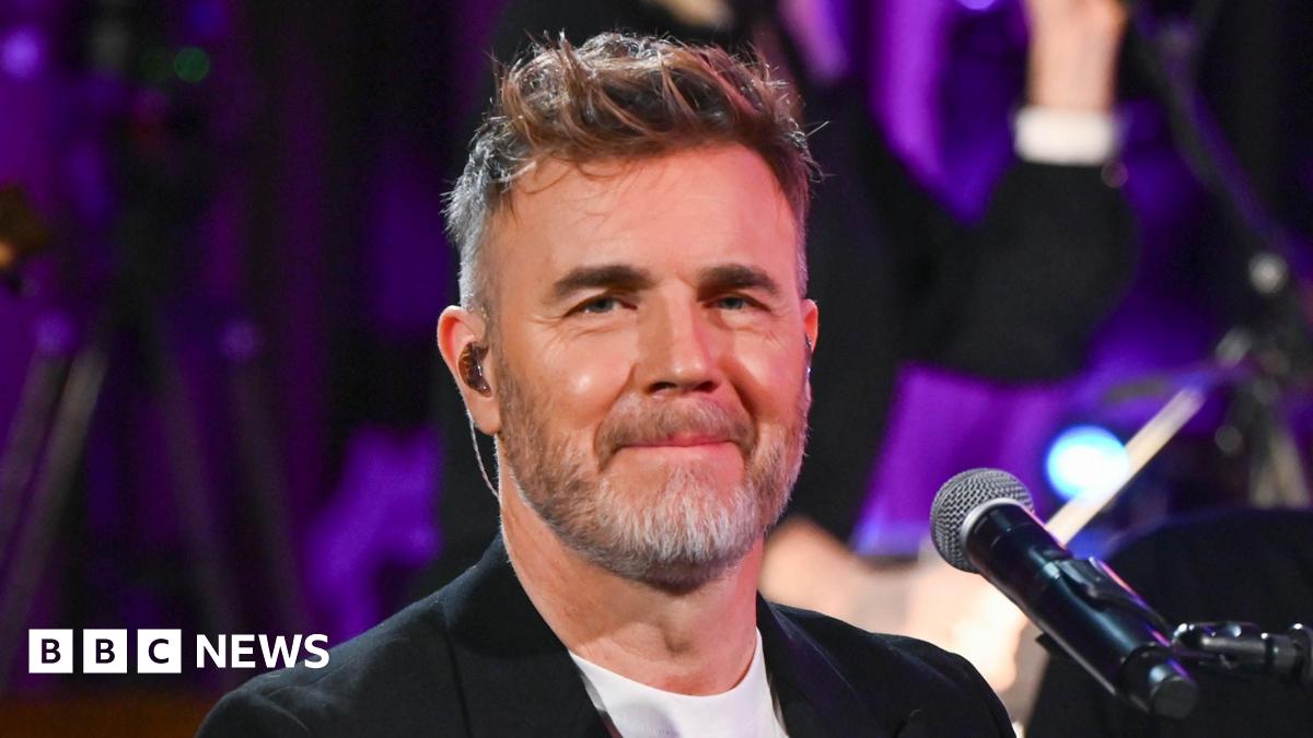 Headshot of Barlow in front of a mic. He wears an in-ear monitor. You can see the top of his white t-shirt and black blazer.