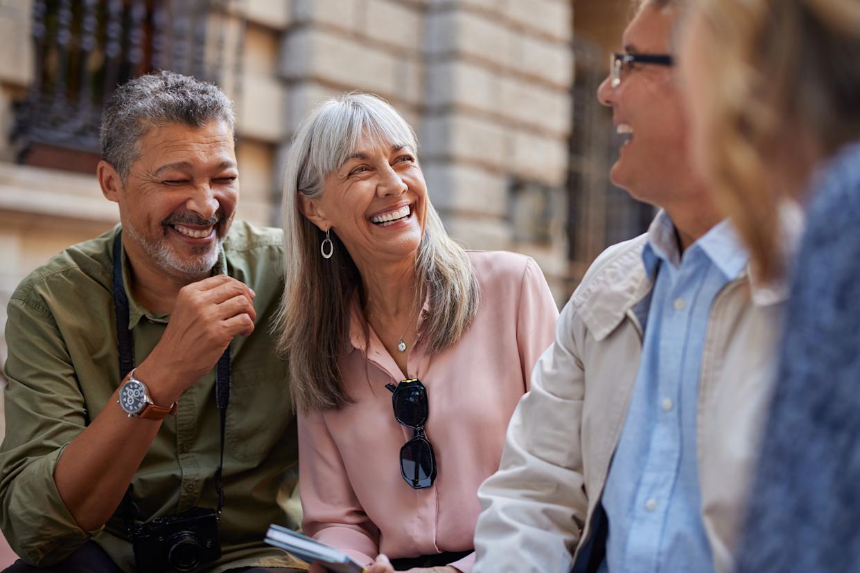 Older friends sit and laugh together while talking during a day out. Group of senior men and women enjoying a cheerful conversation in a city street. Close up of happy mature multiethnic people sharing a fun moment outdoor.