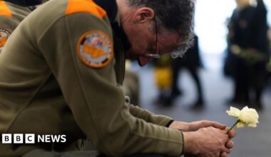 A bespectacled member of the civil protection unit wearing a khaki uniform, squats as he holds a white rose with both hands, with his head lowered