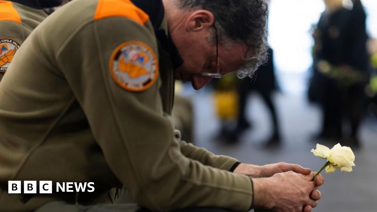 A bespectacled member of the civil protection unit wearing a khaki uniform, squats as he holds a white rose with both hands, with his head lowered
