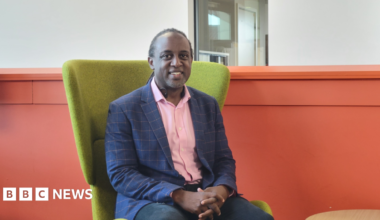 Shows a man in a pink shirt, blue and pink jacket and dark trousers sitting in a green chair in front of an orange wall.