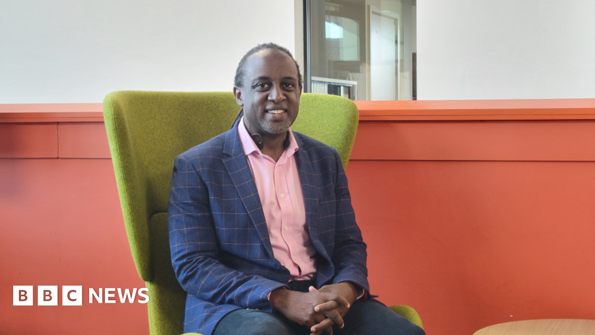 Shows a man in a pink shirt, blue and pink jacket and dark trousers sitting in a green chair in front of an orange wall.