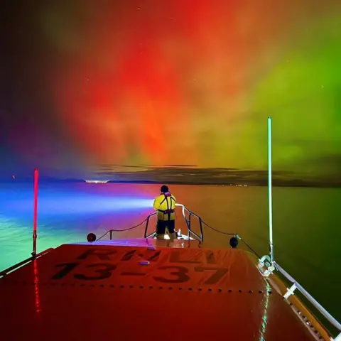 Michael MacDonald/Invergordon RNLI A lifeboat volunteers looks out from Invergordon's lifeboat across the sea towards a red and green aurora.