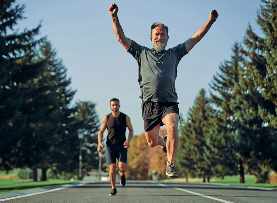 Two people running on a road lined with trees -- the younger person is trailing behind the older person who has leaped into the air with arms raised