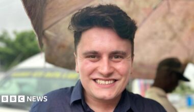 A young man is centre frame and smiling to camera.  He has dark hair and is wearing a navy blue shirt.