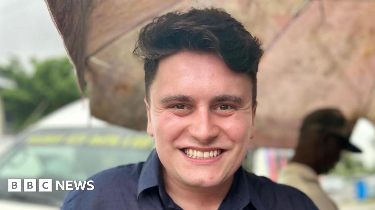 A young man is centre frame and smiling to camera.  He has dark hair and is wearing a navy blue shirt.