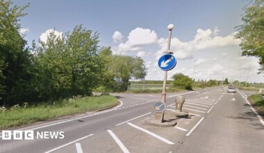 The junction of a main raid with two side roads, surrounded by trees and green trees. The grey asphalt is split by a crossing point with blue signs telling motorists to keep left. White road markings denote the turn-offs.