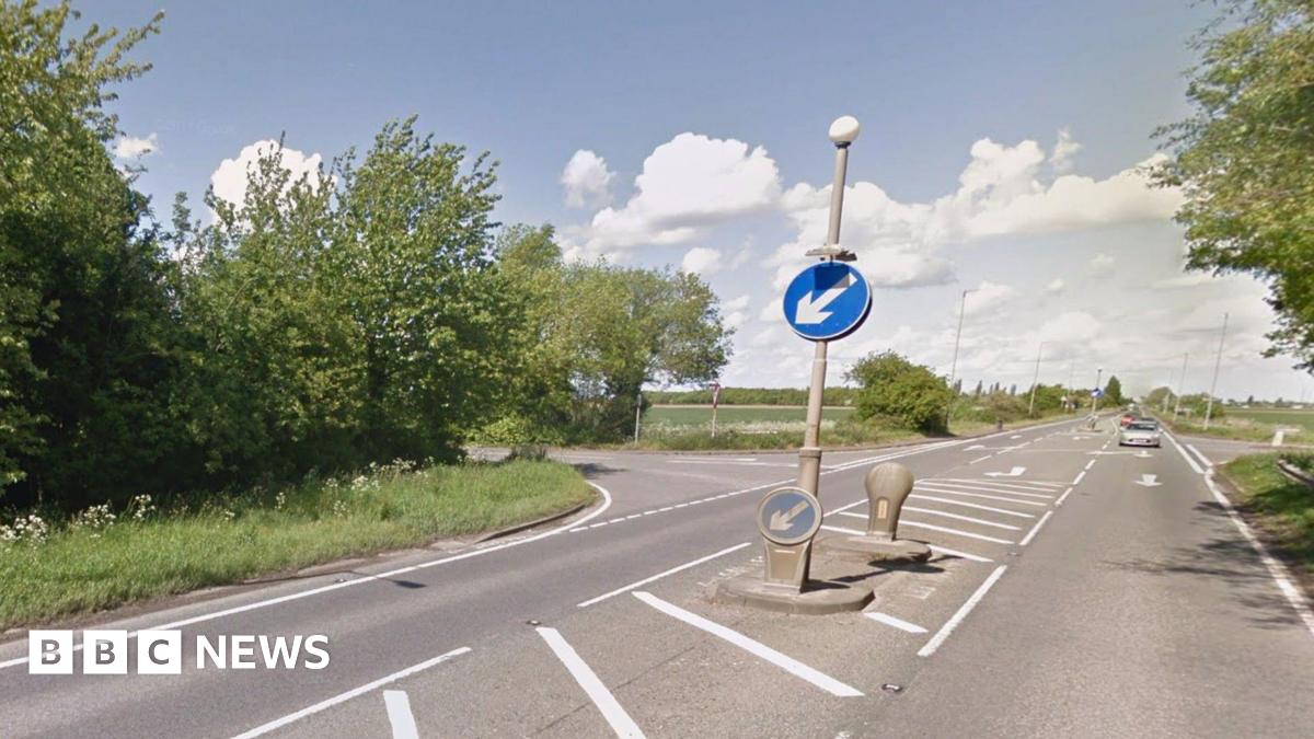 The junction of a main raid with two side roads, surrounded by trees and green trees. The grey asphalt is split by a crossing point with blue signs telling motorists to keep left. White road markings denote the turn-offs.